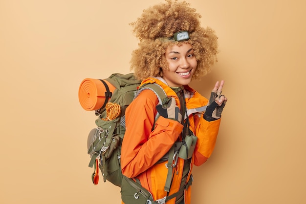 Cheerful young female camper dressed in orange jacket makes peace gesture enjoys active lifestyle carries backpack being in good mood isolated over beige background Hiking and traveling concept