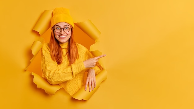 Free photo cheerful young female model has red hair toothy smile pointing at copy space, dressed in yellow sweater jumper eyeglasses.