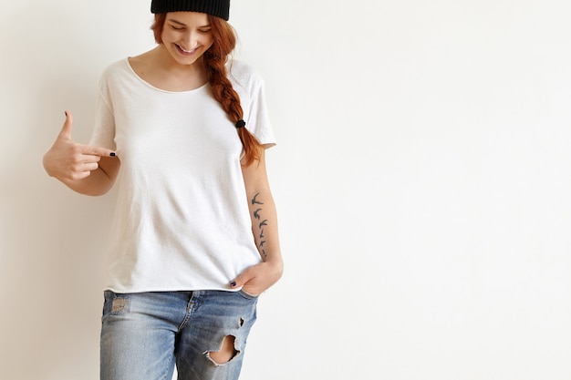 Free Photo cheerful young female with ginger hair and tattoo looking down and pointing index finger at her white oversize t-shirt