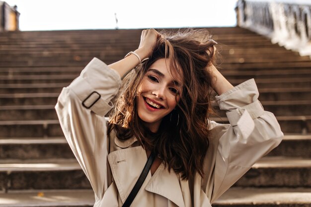 Cheerful young girl with dark wavy hairstyle and red lips, wearing beige trench, smiling, messing up hair and looking at front against wall of old city stairs