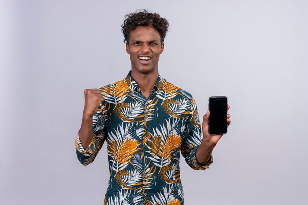 Cheerful young good-looking dark-skinned man with curly hair in leaves printed shirt holding smartphone with clenching fist 