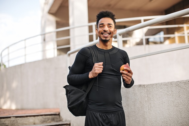 Cheerful young guy in black long-sleeved t-shirt and shorts smiles widely, holds backpack and apple, poses outside