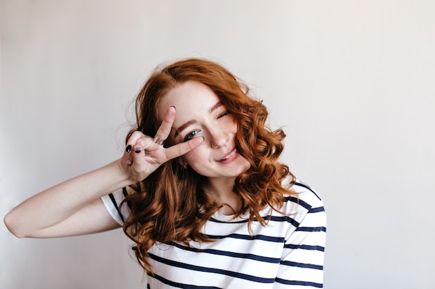 Free Photo cheerful young lady with pale skin and red hair posing with peace sign. amazing girl in striped t-shirt isolated.