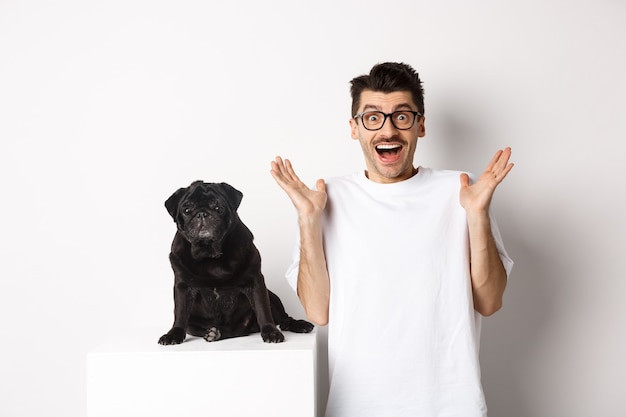 Free photo cheerful young man in glasses standing with his pet, rejoicing and staring at camera amused, hear great news, standing with pug over white background