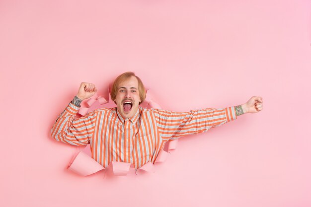 Cheerful young man poses in torn coral paper hole wall emotional and expressive