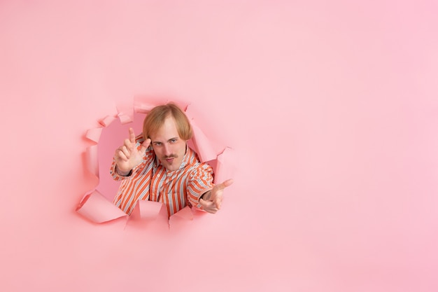 Cheerful young man poses in torn coral paper hole wall emotional and expressive