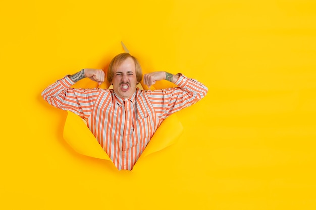 Free photo cheerful young man poses in torn yellow paper hole wall emotional and expressive