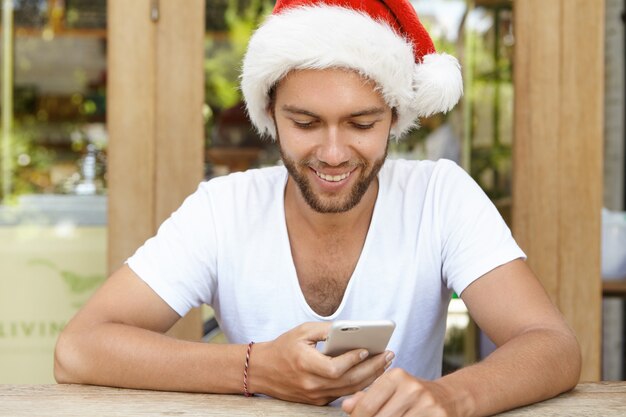 Cheerful young man wearing Santa Claus hat greeting his family on New Year's Day, texting messages online on mobile phone.