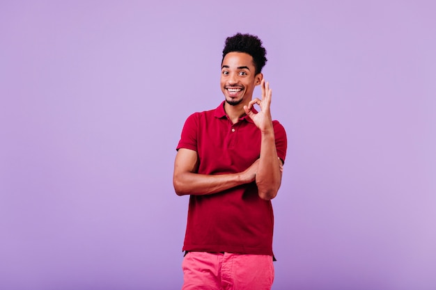 Cheerful young man wears red t-shirt posing with okay sign. funny african male model isolated.