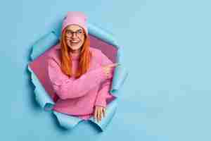 Free photo cheerful young redhead woman wears pink hat and knitted sweater pointing at copy space from paper hole