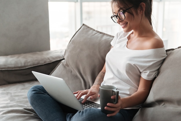 Cheerful young woman holding cup of coffee using laptop computer.