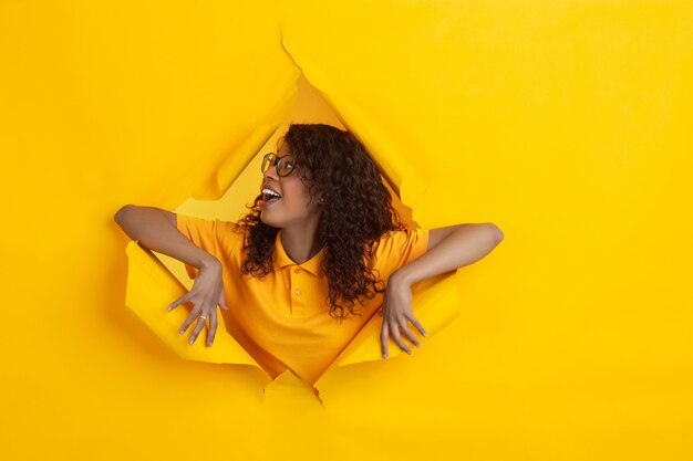 Cheerful young woman poses in torn yellow paper hole background, emotional and expressive