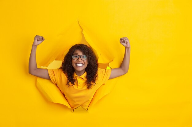 Cheerful young woman poses in torn yellow paper hole background, emotional and expressive