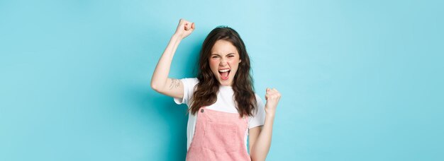 Cheerful young woman rooting for team watching sports game and cheering raising hand up in fist pump gesture shouting to encourage person standing over blue background