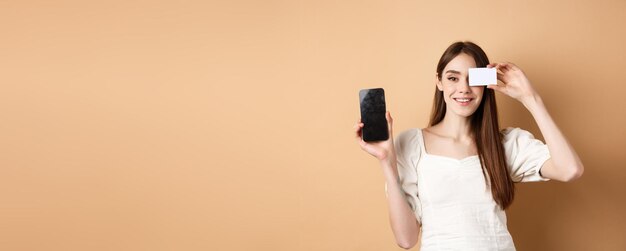 Free photo cheerful young woman showing plastic credit card and empty mobile phone screen smiling pleased at ca