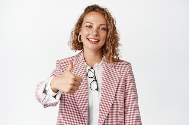 Free Photo cheerful young woman showing thumbs up, approve something good, smiling and nod, standing in suit against white background