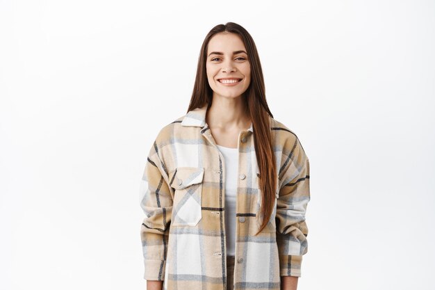 Cheerful young woman in stylish oversize shirt smiling, showing white perfect smile, looking happy at camera, standing over white background