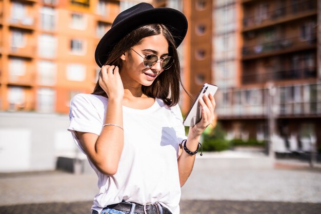 Cheerful young woman wearing black hat in city street typing a message. 