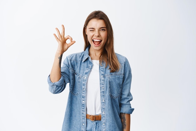 Free Photo cheerful young woman winking and showing ok sign say alright or yes approve something good recommending product and quality standing pleased against white background