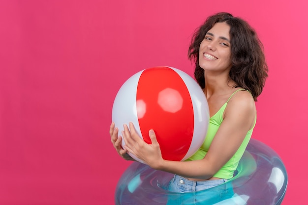 A cheerful young woman with short hair in green crop top smiling and holding inflatable ball 