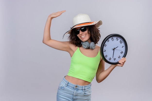 A cheerful young woman with short hair in green crop top wearing sunglasses and sun hat holding a wall clock and raising hand on a white background