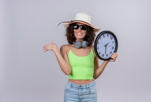 A cheerful young woman with short hair in green crop top wearing sunglasses and sun hat holding a wall clock and showing goodbye on a white background