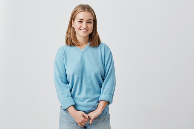 Cheerfulcharming young woman wearing blue sweater with fair straight hair smiling happily while receiving some positive news. Pretty girl dressed in blue looking  with joyful smile.