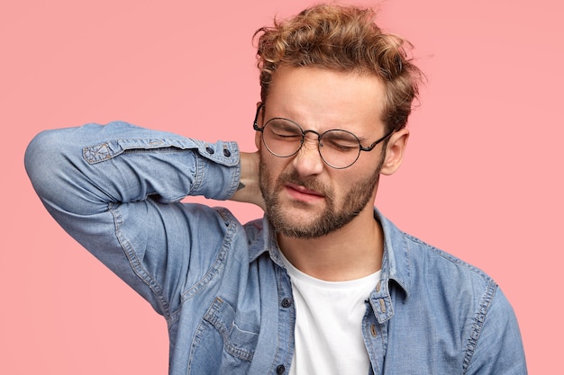 Cheerless man gets stiff neck, suffers from pain as has sedentary lifestyle and works for long time at computer, frowns face in dissatisfaction, wears spectacles and denim shirt, stands indoor