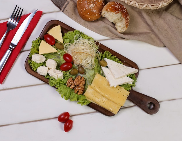 Cheese platter with tomatoes, nuts and olives with cutlery and bread buns around.