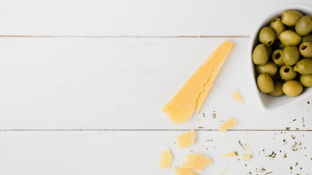 Cheese slice with green olives in the bowl on white wooden desk