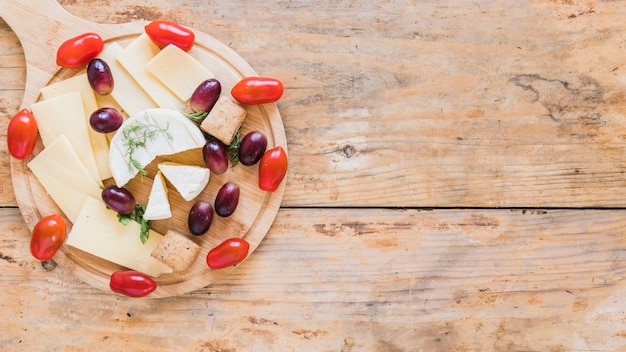 Cheese slices and blocks served with grapes and cherry tomatoes on chopping board over the desk