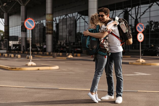 Cheetful man in sunglasses, white tee, jeans hugs with blonde woman in plaid shirt