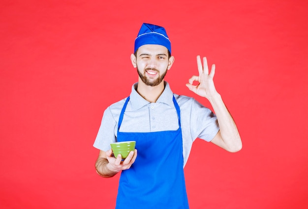 Chef in blue apron holding a green ceramic cup and showing satisfaction sign. 