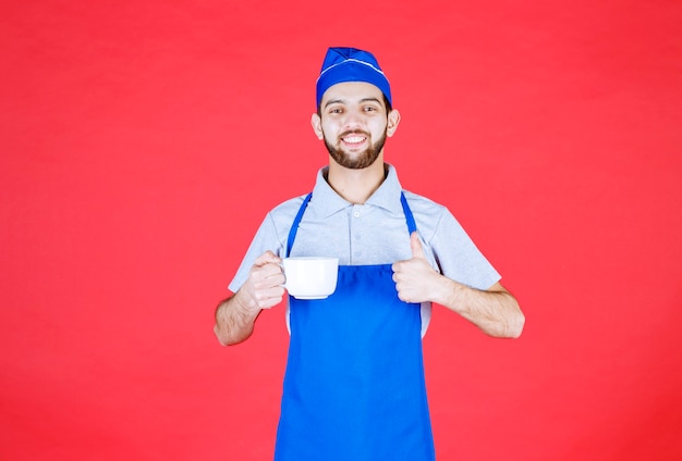 Chef in blue apron holding a white ceramic cup and showing enjoyment sign. 