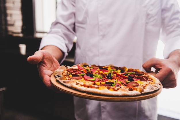 Free photo chef holding a wooden plate with a delicious pizza on it in the kitchen