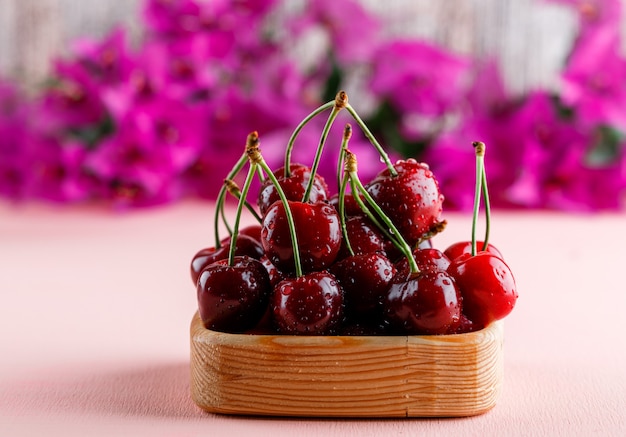 Cherries with flowers in a wooden plate on pink surface, side view