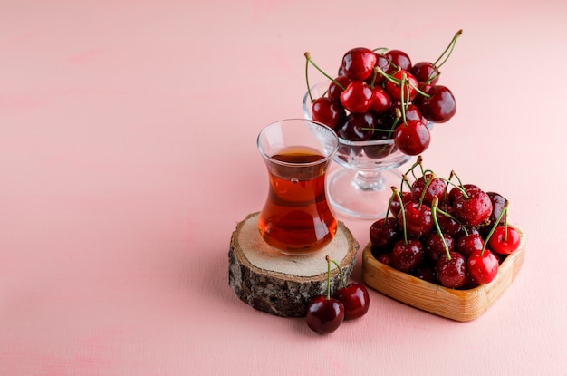 Free Photo cherries with glass of tea on wooden board in wooden plate and vase on pink surface