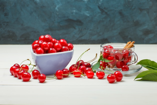 Cherries with leaves, spices in bowl and mug on wooden and dark blue, side view.