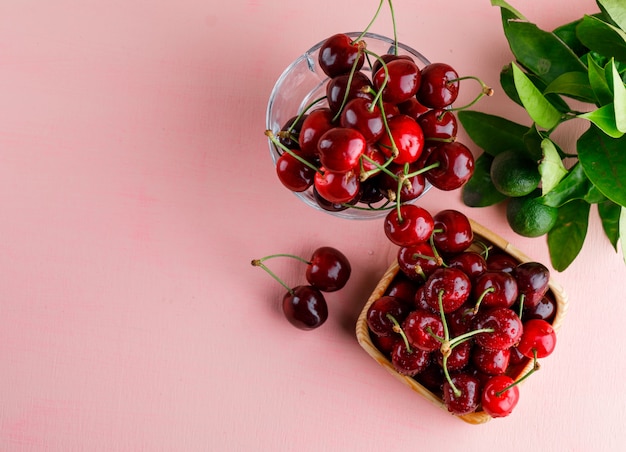 Cherries in wooden plate and vase with lemons and leaves top view on a pink surface