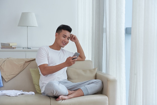 Chherful guy enjoying his free time checking social media seated comfortably on the sofa