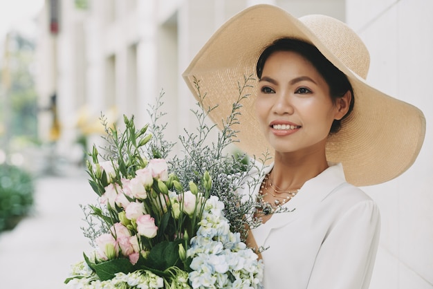 Chic wealthy Asian woman in large straw hat posing in street with fresh flower bouquet