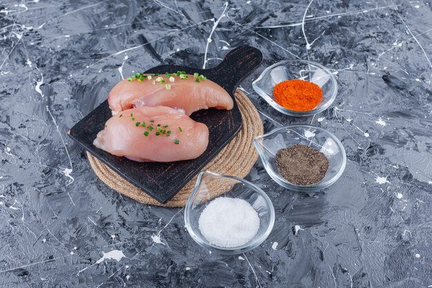 Chicken breast on a cutting board on a trivet next to bowls full of spices, on the blue table.