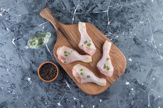 Chicken drumsticks on a cutting board next to oil and spice bowls , on the blue surface.