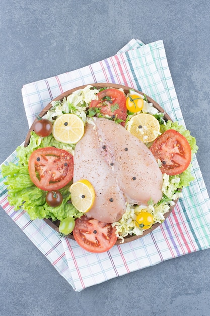 Chicken fillets and sliced vegetables on wooden plate.