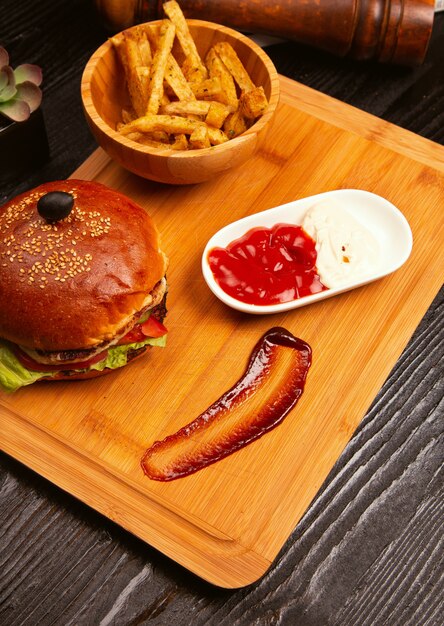 Chicken meat burger with tomato and lettuce inside and french fries served with black olive and ketchup on a wooden tray