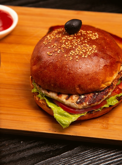 Chicken meat burger with tomato and lettuce inside served with black olive and ketchup on a wooden tray.