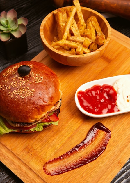 Chicken meat burger with tomato slices and lettuce served with french fries, ketchup and mayonnaise on wooden board