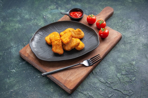 Chicken nuggets on a black plate and fork on wooden board tomatoes ketchup on dark surface with free space