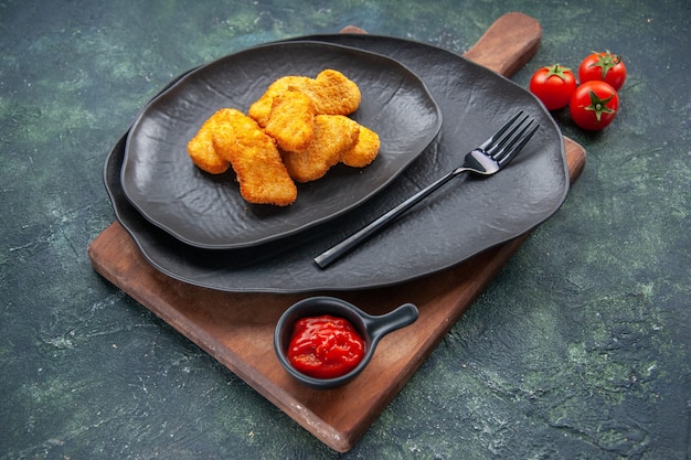 Chicken nuggets on a black plate and fork on wooden board tomatoes ketchup on dark surface