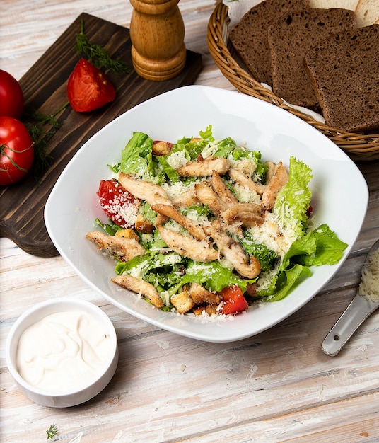 Chicken parmesan caesar salad with lettuce, cherry tomatoes inside a white bowl, served with sauce and bread.